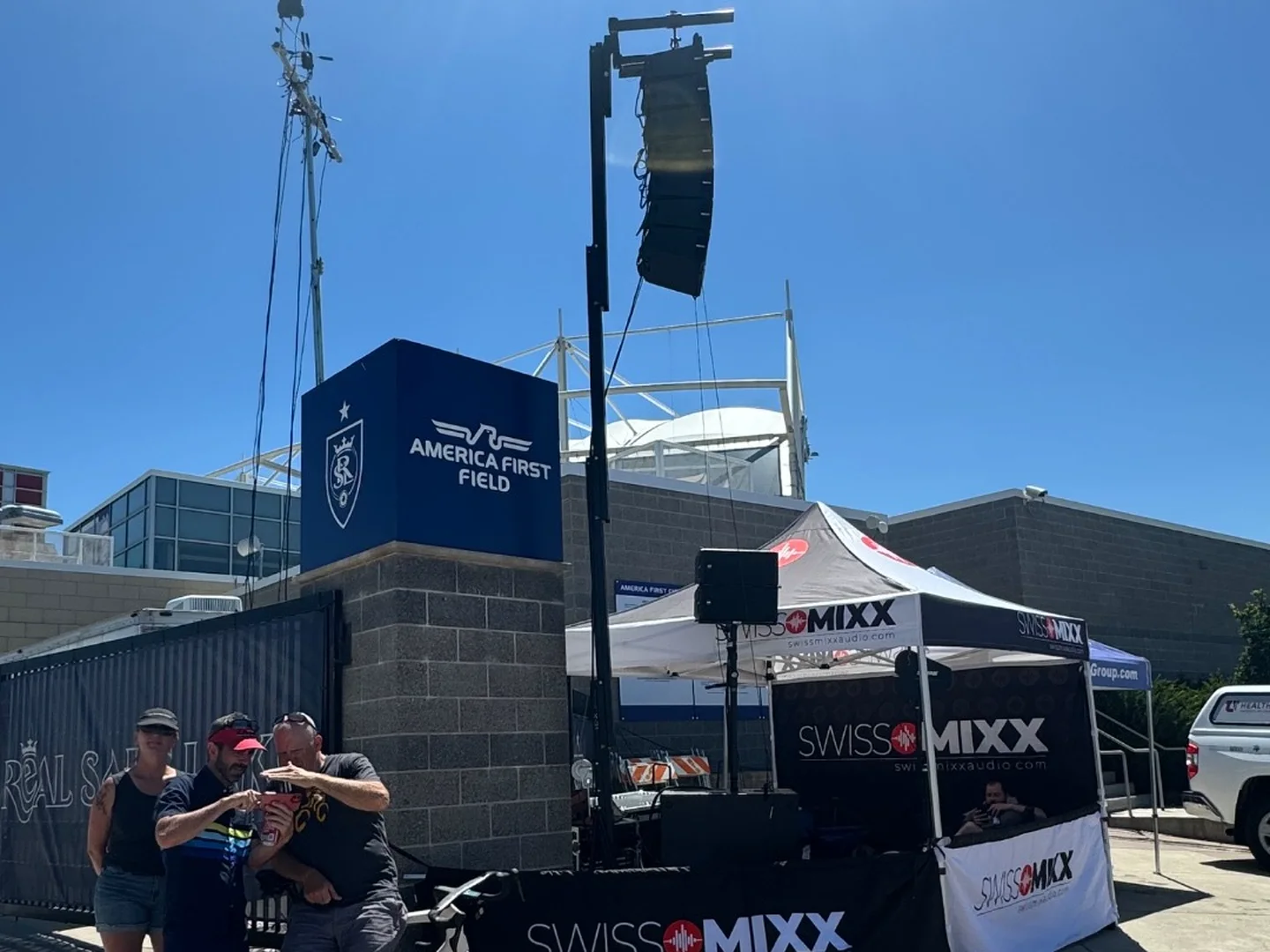 SwissMixx Audio line array flown on a lift beside the SwissMixx tent at a criterium outside America First Field, with riders in the foreground