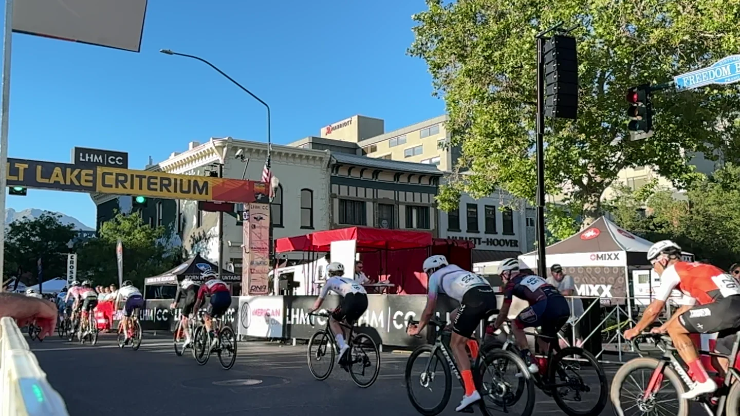 Criterium field racing past the Salt Lake Criterium arch on a closed city street, with vendor tents and the SwissMixx Audio tent along the course